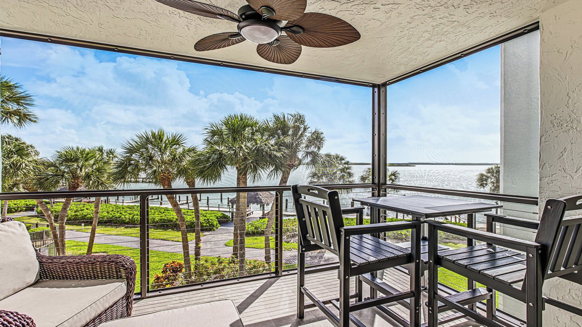 A balcony with a ceiling fan, a table, chairs, and a view of a water body and palm trees.