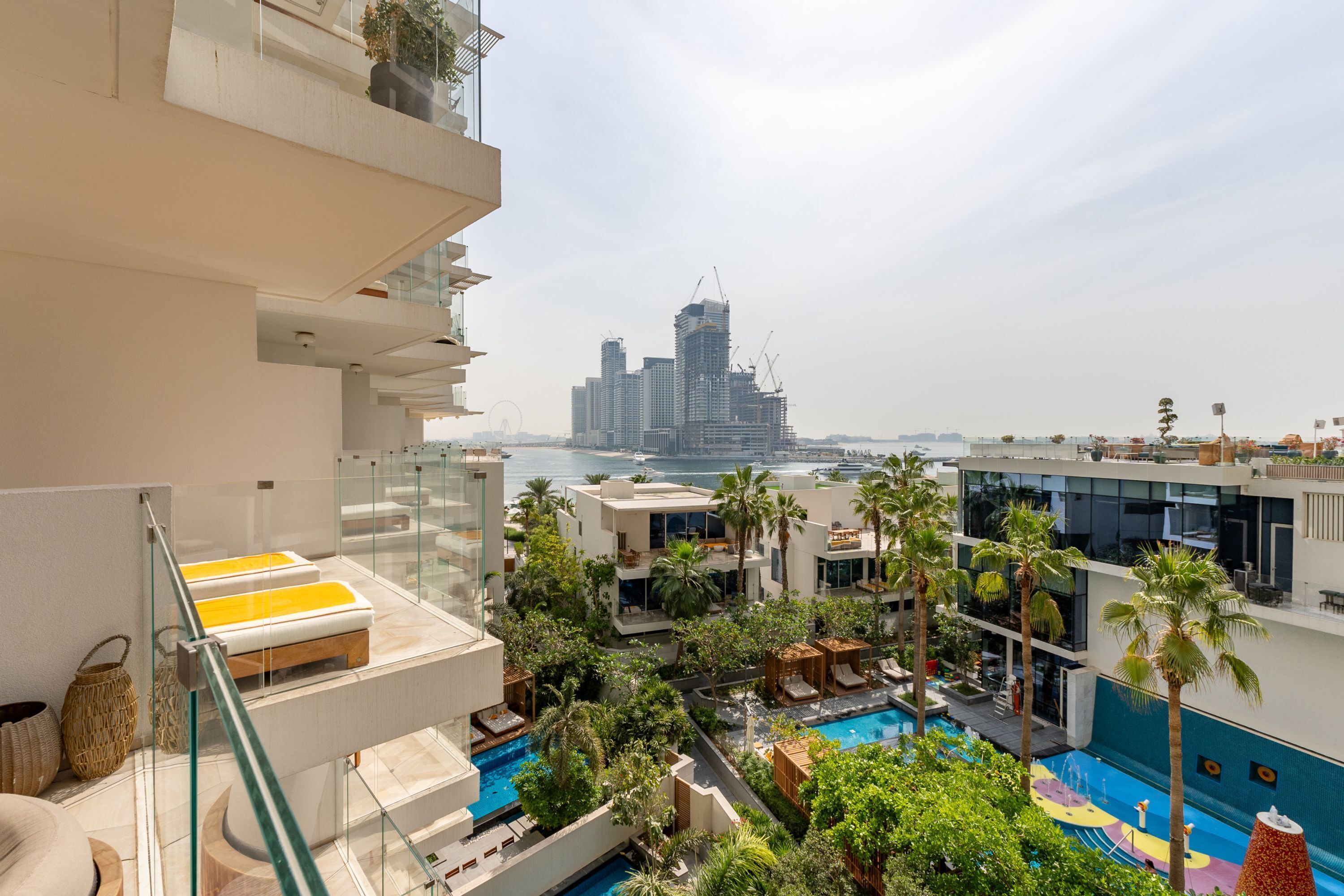 A rooftop pool area with multiple pools, palm trees, and a view of a city skyline with high-rise buildings.
