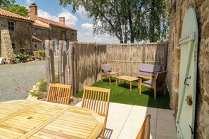 A patio with a wooden table and chairs, a small garden area with artificial grass, and a stone building in the background. - A charming vacation rental with a pool, nestled in the peaceful Vendée countryside. (Saint-Juire-Champgillon)