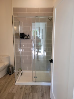 A modern bathroom with a glass shower enclosure, a toilet, and a trash bin. - New Hidden Creek Studio (Ucluelet)