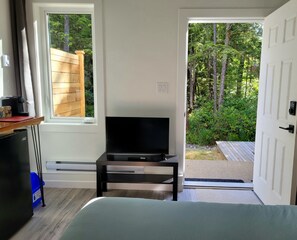 A room with a bed, a TV on a stand, a window, and a door leading to an outdoor area with greenery. - New Hidden Creek Studio (Ucluelet)