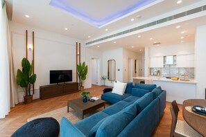 Una sala de estar moderna con un sofá grande, una mesa de centro y un televisor de pantalla plana. Se observa un área de cocina con armarios y una encimera.