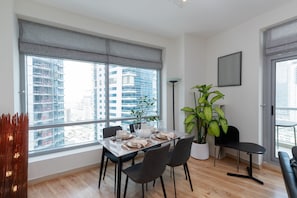 Un comedor con una mesa dispuesta para cuatro, ventanas amplias con persianas, una planta en maceta y una lámpara moderna.