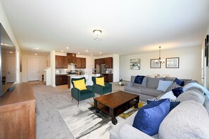 A modern living room with a sofa, coffee table, and chairs, a kitchen in the background, and a dining area.