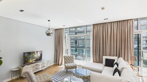 Ein modernes Wohnzimmer mit Sofa, Sessel und Couchtisch, großen Fenstern mit Vorhängen sowie einem Flachbildfernseher auf einer Anrichte.