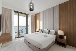 Un hotel moderno con una cama grande, una mesita de noche con lámpara, vista al mar y un balcón con una silla.