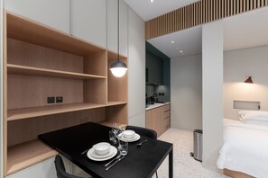 Ein modernes Hotelzimmer mit Esstisch für zwei Personen, einem Bett mit weißer Bettwäsche und einer kleinen Küche im Hintergrund.