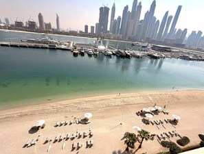 Eine Küstenstadt mit einem Yachthafen, Hochhäusern und einem Sandstrand mit Liegestühlen und Sonnenschirmen.