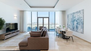 A modern living room with a large window, a sofa, a dining table, and a TV. - Grand 3BR Beachfront Living | Palm Jumeirah (Dubai)