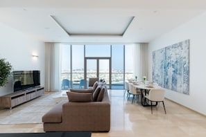 A modern living room with a large window, a sofa, a dining table, and a TV. - Grand 3BR Beachfront Living | Palm Jumeirah (Dubai)