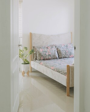 Een bed met bloemenbedekking, een potplant en een houten stoel in een kamer met witte wanden en een betegelde vloer.