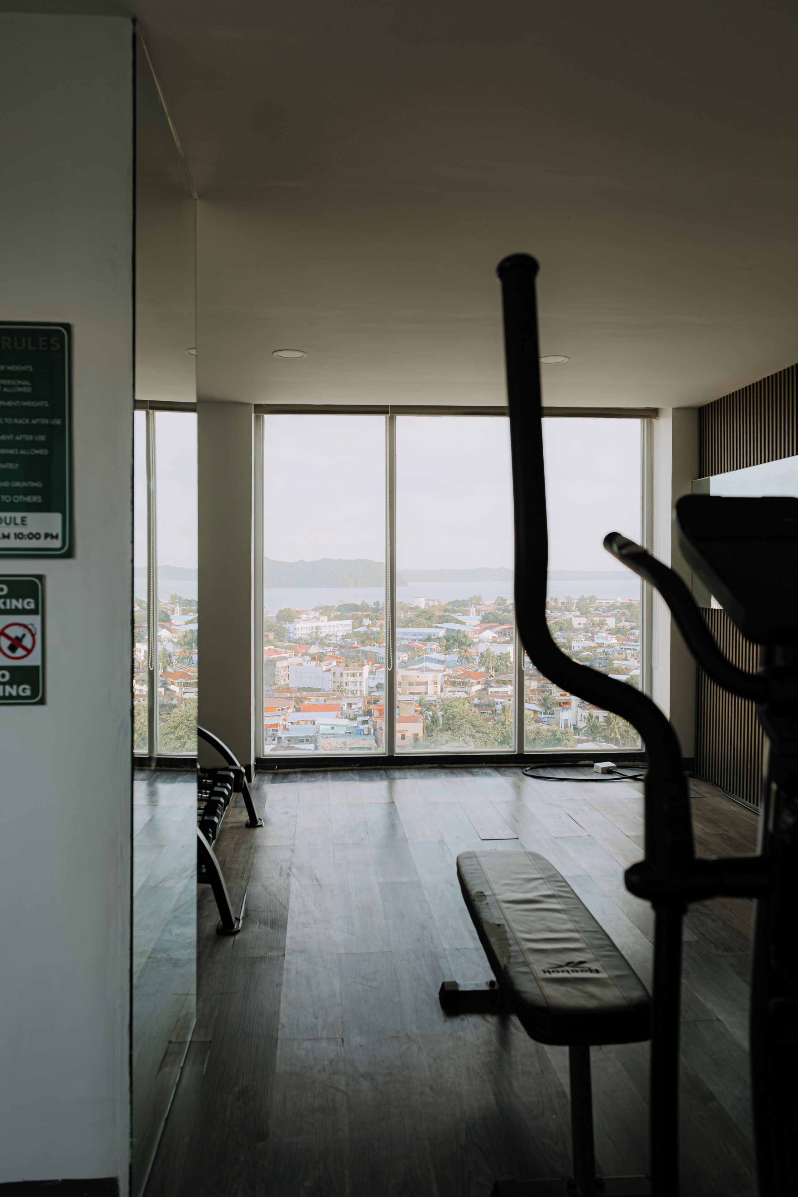 A gym with a bench and exercise equipment, large windows with a city view, and a sign with rules.