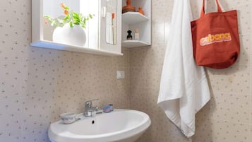 Une salle de bain avec un lavabo blanc, un miroir au-dessus et une serviette accrochée au mur. Un sac rouge est suspendu au porte-serviettes.