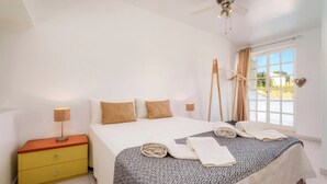 Een slaapkamer met een bed, bedlampen, een houten nachtkastje, een raam met gordijnen en een plafondventilator.