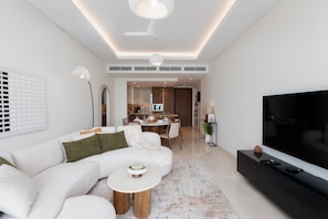 A modern living room with a sectional sofa, a round coffee table, a flat-screen TV, and a dining area in the background. - Urban 3BR Apt | Walk to Dubai Mall & Opera (Dubai)