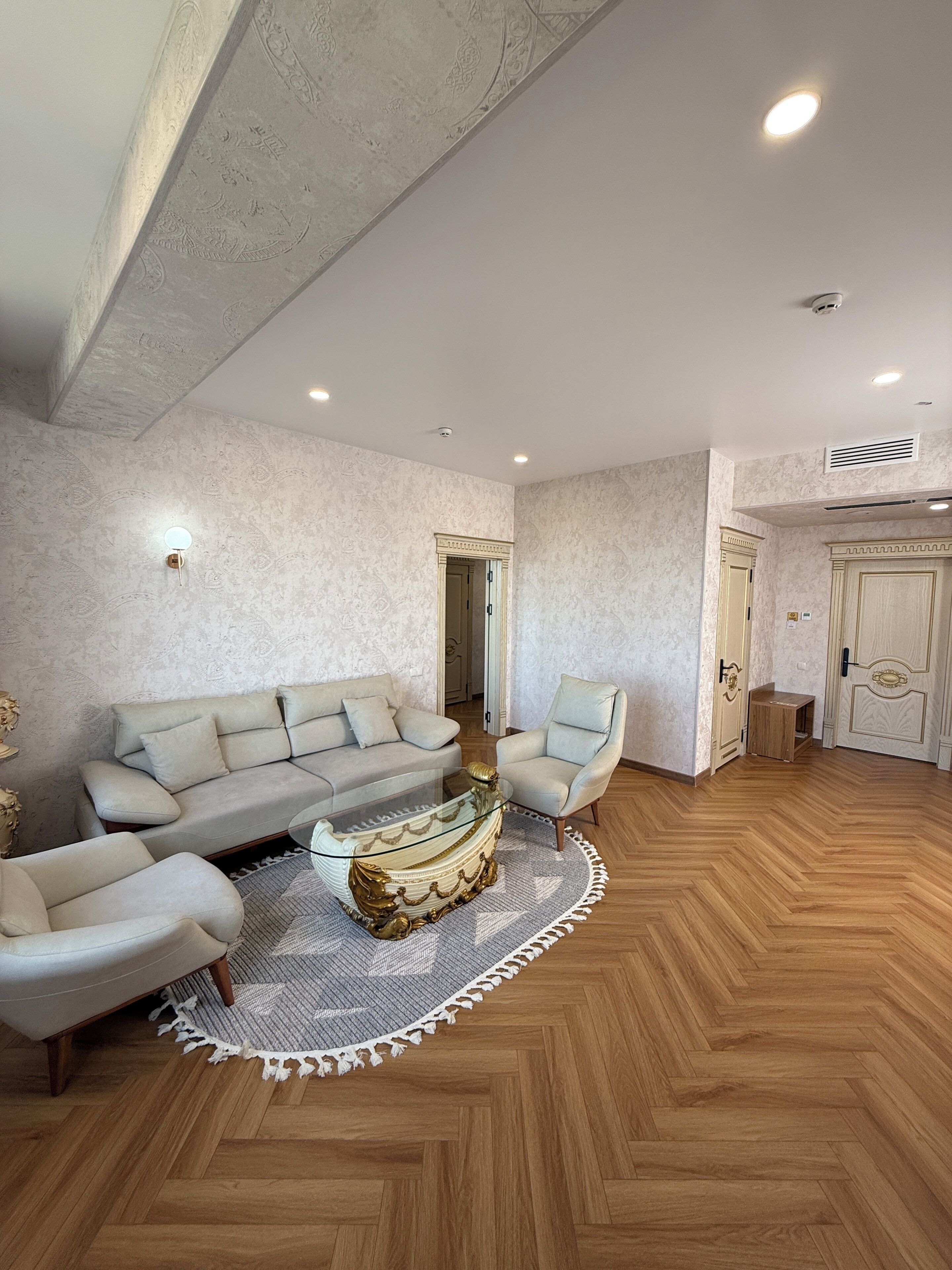Un ampio soggiorno con pavimento in legno a spina di pesce, un divano color beige, un tavolino in vetro e una poltrona dai toni chiari.