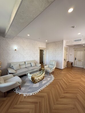 A spacious living room with a herringbone wood floor, a beige sofa, a glass coffee table, and a light-colored armchair. - Safia Plaza Hotel (Bukhara)
