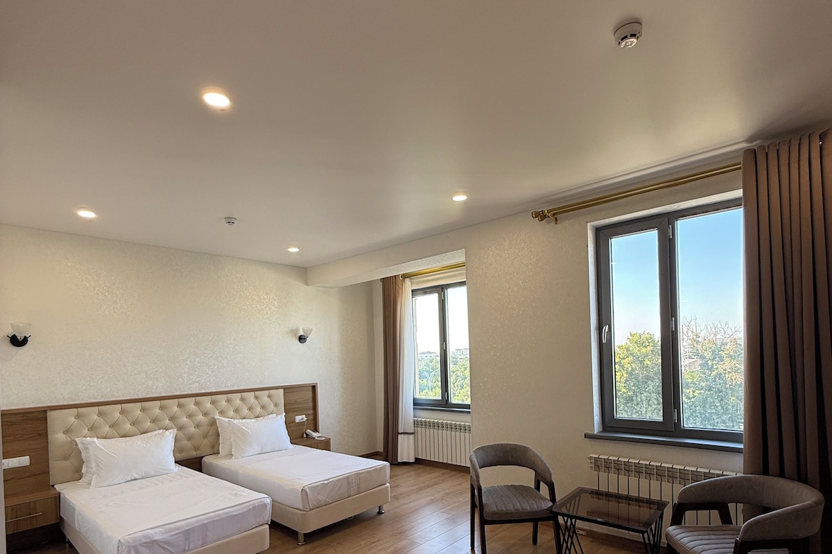 A hotel room with two beds, a small table, a chair, a window with curtains, and a radiator.