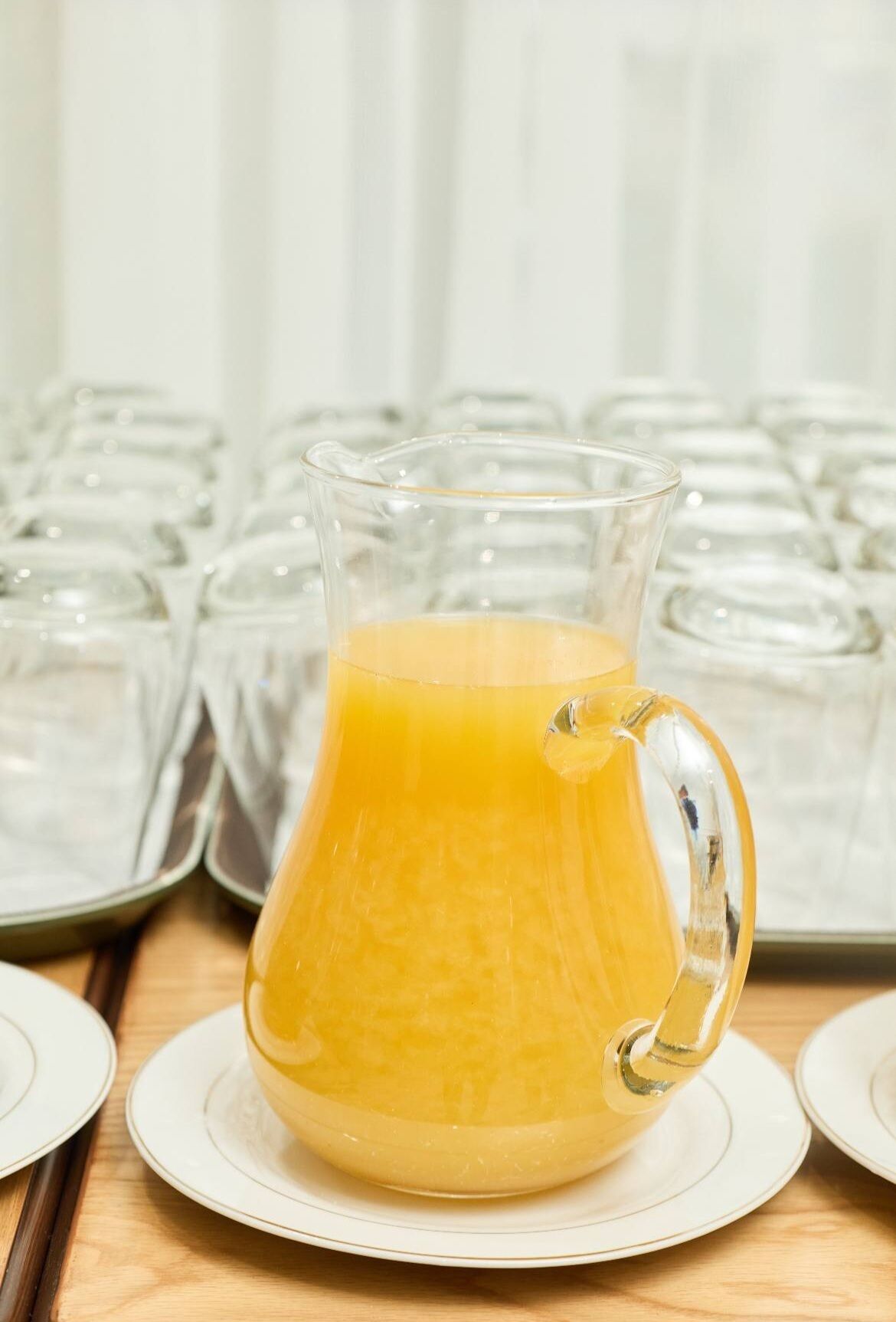 A pitcher of orange juice on a plate with a tray of glasses in the background.