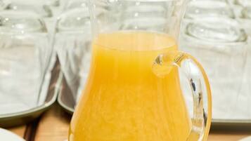 A pitcher of orange juice on a plate with a tray of glasses in the background.