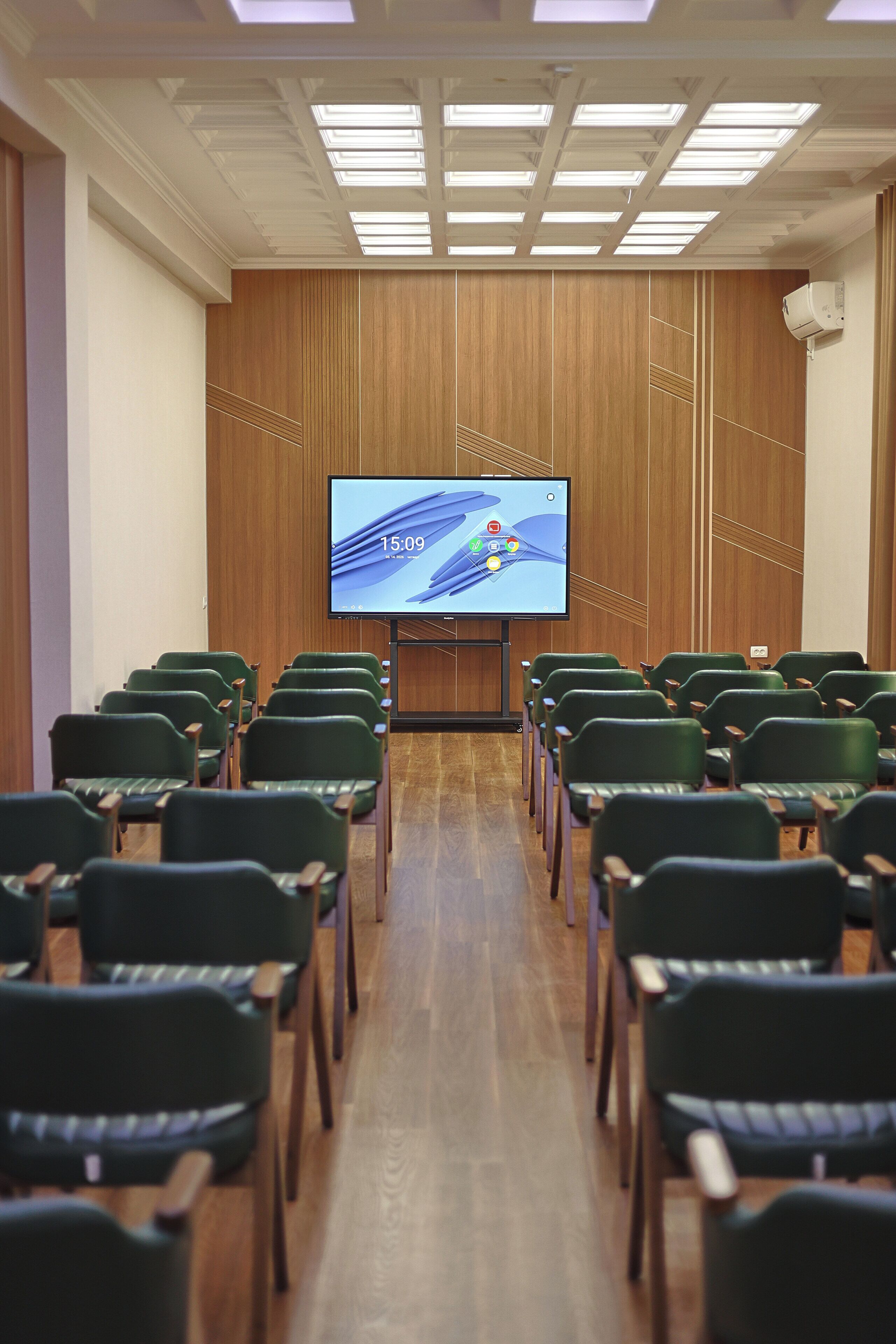 A conference room with rows of green chairs, a large screen, and wooden paneling on the walls.