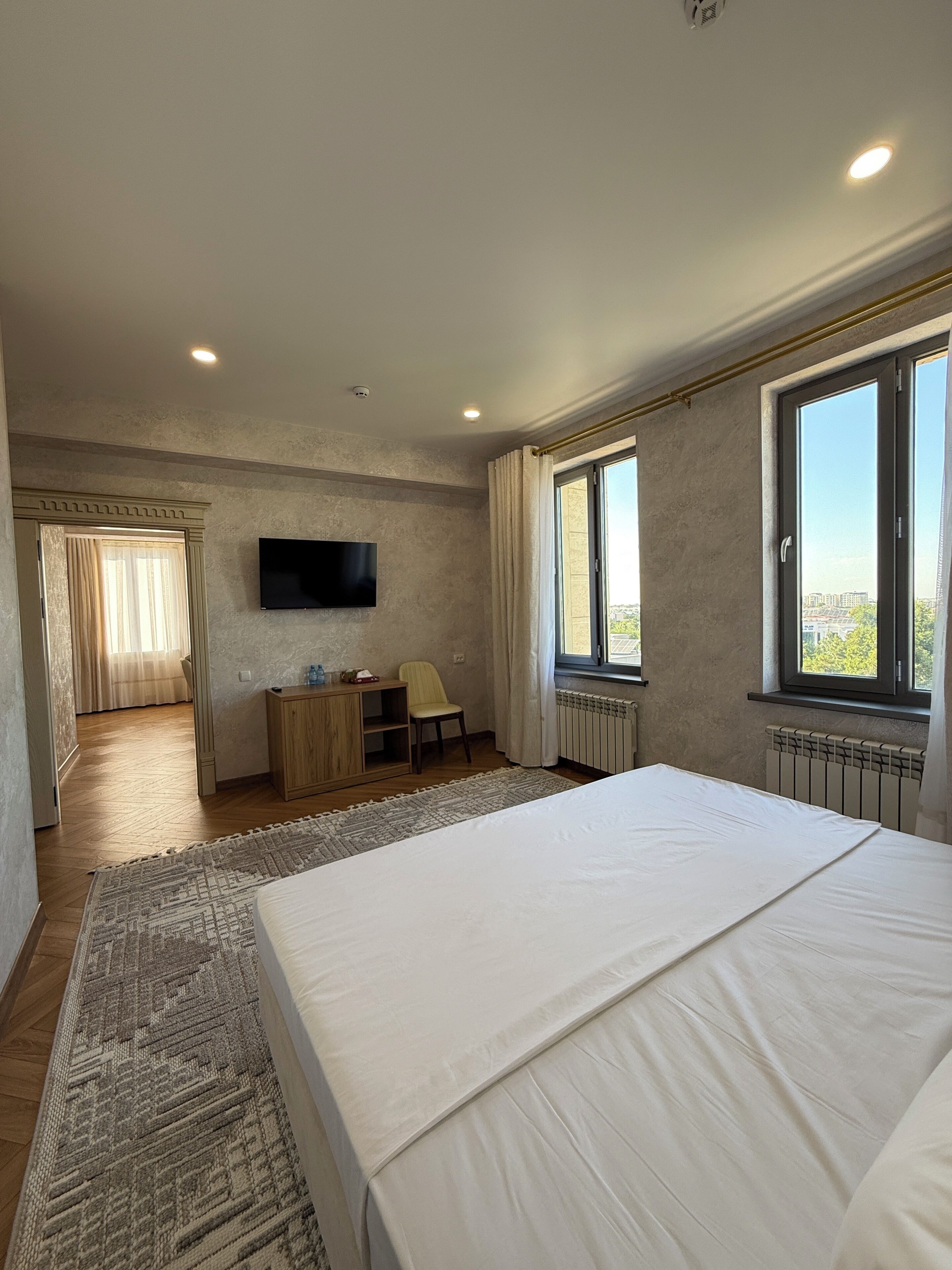 Ein Schlafzimmer mit Bett, Schreibtisch, Stuhl, Fernseher und großen Fenstern.