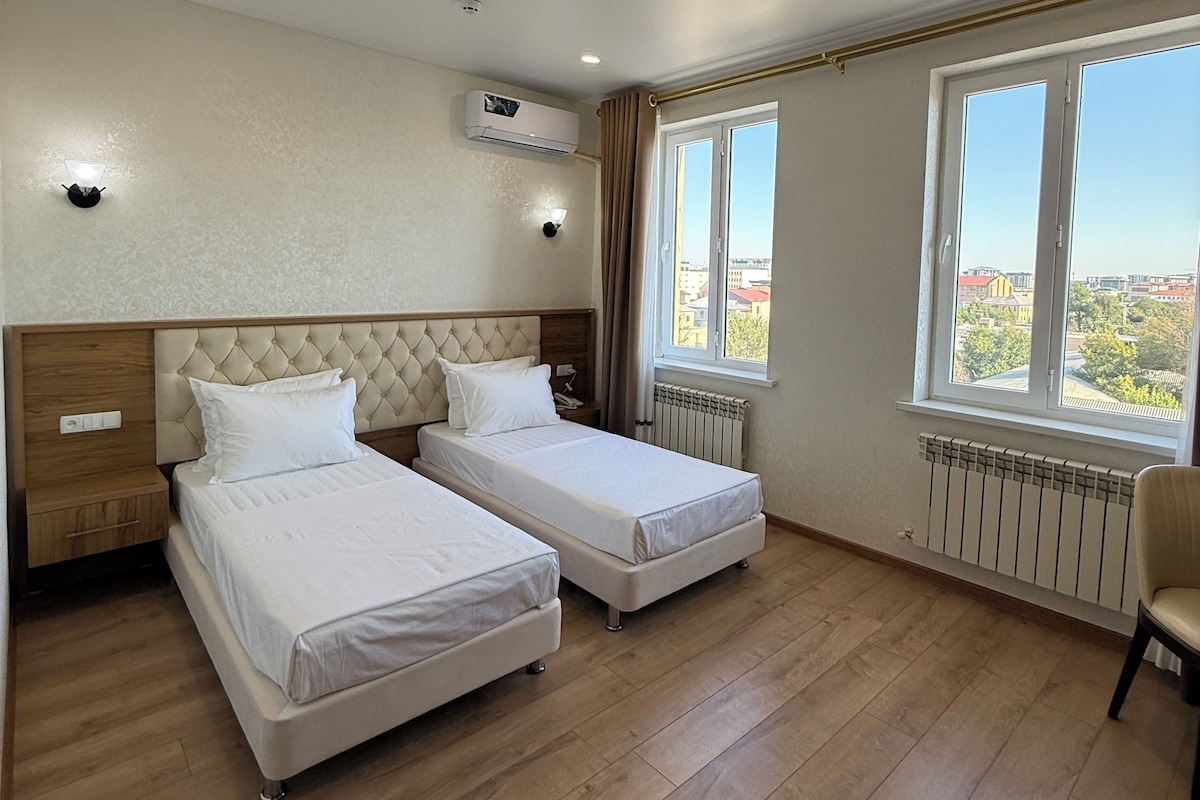 A hotel room with two beds, a window with a view, an air conditioning unit, and a radiator.