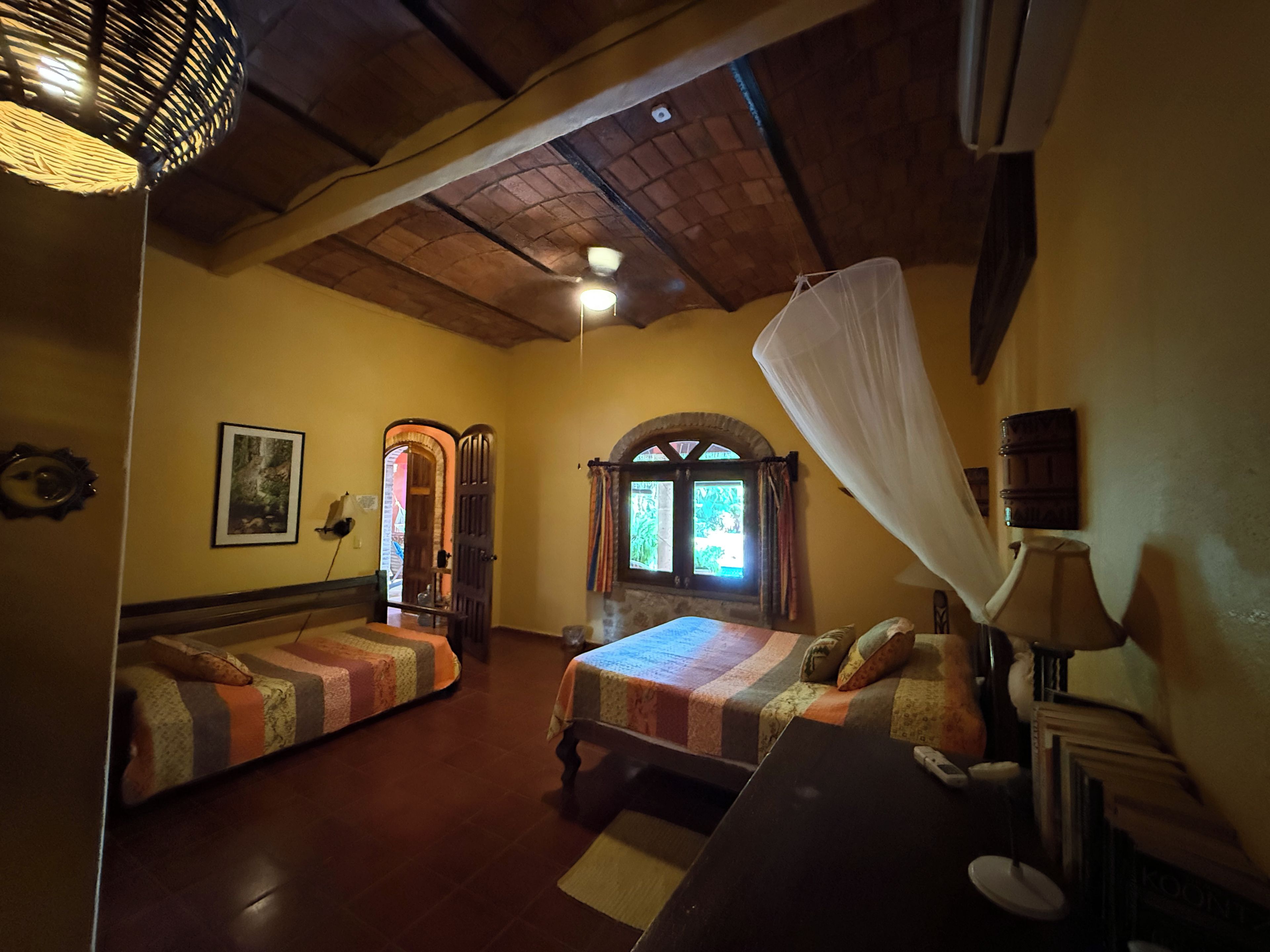 A room with two beds, a wooden ceiling, a window with curtains, and a wall-mounted lamp.