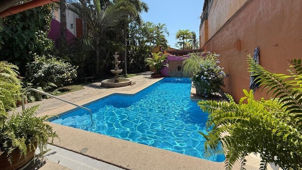 A swimming pool with a tiled deck, surrounded by greenery and a building with a red wall. - Casa Sorpresa mexican charm by the beach (Sayulita)