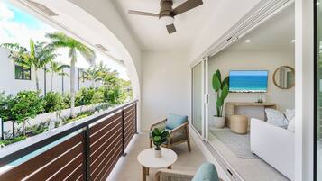 A balcony with a view of palm trees and a pool, a ceiling fan, a sofa, and a television.