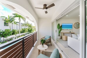 A balcony with a view of palm trees and a pool, a ceiling fan, a sofa, and a television. - The Palmetto: Boca Pool Suite + Beach (Boca Raton)