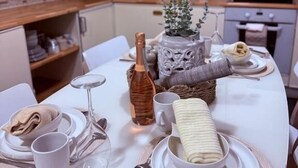 Ein ordentlich gedeckter Esstisch mit Tellern, Gläsern und einer Flasche, vor einer Küchenkulisse mit Schränken und Geräten.