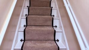 Eine Treppe mit beigen Teppichstufen, weißen Geländern und einer Pflanze oben.