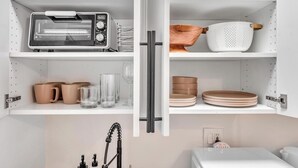 Une cuisine équipée d’un grille-pain encastré, d’une bouilloire et d’une machine à café. On y trouve des étagères avec des tasses, des verres et des assiettes.