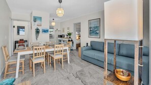 Un salon moderne avec un canapé bleu, une table à manger en bois et des chaises. On aperçoit à l’arrière-plan un coin cuisine avec un micro-ondes et une plante.