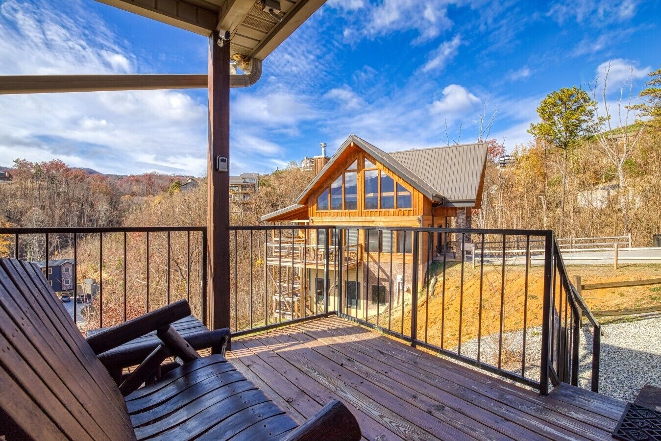 Una terraza de madera con mobiliario exterior, un espacio cubierto y vistas a una cabaña con un gran ventanal y techo inclinado.