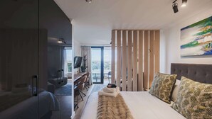 A modern hotel room with a large bed, a desk, a TV, and a balcony with a view.