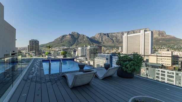 Ein Pool auf dem Dach mit Liegestühlen, einer Glasbrüstung und einer Stadtansicht mit Bergen im Hintergrund.