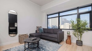 Ein modernes Wohnzimmer mit einem Ecksofa, einem runden Couchtisch, einem großen Fenster mit Stadtblick, einem Spiegel und einer Topfpflanze.