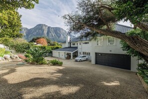 A two-story house with a garage, surrounded by a gravel driveway, and a large tree in the foreground. - Bishopscourt- Secret Garden hideaway (Cape Town)
