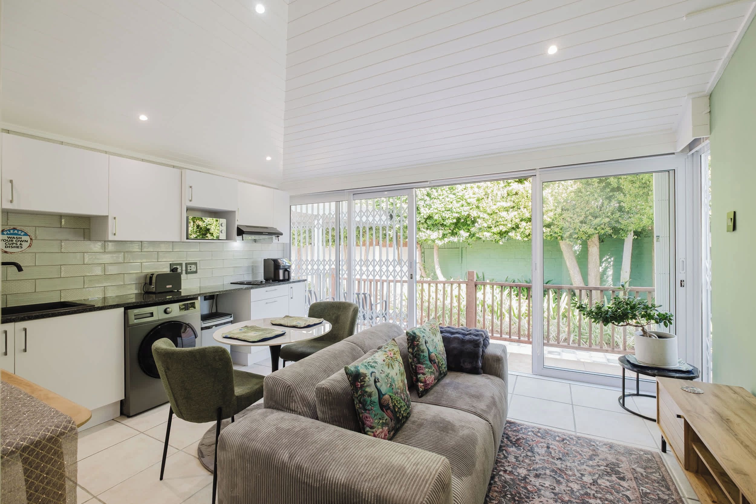 A compact kitchen with a dining area, a sofa, and a view of a green garden through a sliding glass door.