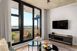 A modern living room with a large sliding glass door leading to a balcony, a flat-screen TV mounted on the wall, a black entertainment unit, and a round glass coffee table. - Riverlands, Sunbird - Nectar Haven (Cape Town)
