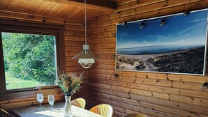A wooden interior with a dining table, chairs, a vase with flowers, and a large window with a view.