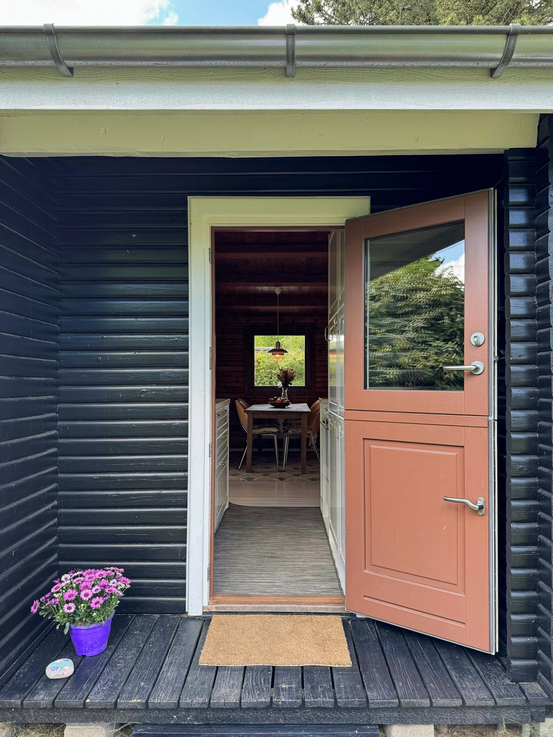 A wooden cabin with a dark exterior, a door leading to a dining area with a table and chairs, and a potted plant on the porch.