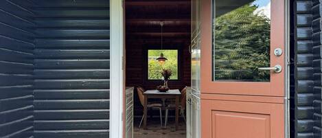 A wooden cabin with a dark exterior, a door leading to a dining area with a table and chairs, and a potted plant on the porch.