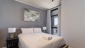 A modern hotel room with a large bed, two bedside lamps, a framed abstract painting, and a large window. - Riverlands- Sunbird Golden Nectar (Cape Town)