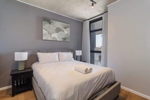 Ein modernes Hotelzimmer mit einem großen Bett, zwei Nachttischlampen, einem gerahmten abstrakten Gemälde und einem großen Fenster.