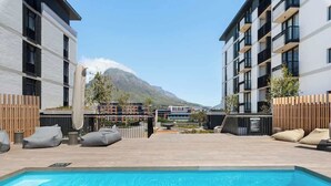 A swimming pool with lounge chairs, a wooden deck, and modern buildings in the background. - Riverlands- Sunbird Golden Nectar (Cape Town)