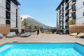 Ein Schwimmbecken mit Liegestühlen, einer Holzterrasse und modernen Gebäuden im Hintergrund.