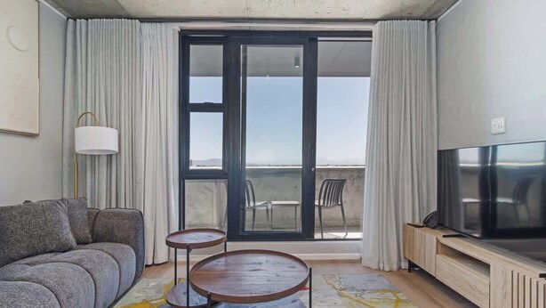 A living room with a sofa, two small tables, a TV, and a view of the sea through a large window. - Riverlands- Sunbird Golden Nectar (Cape Town)